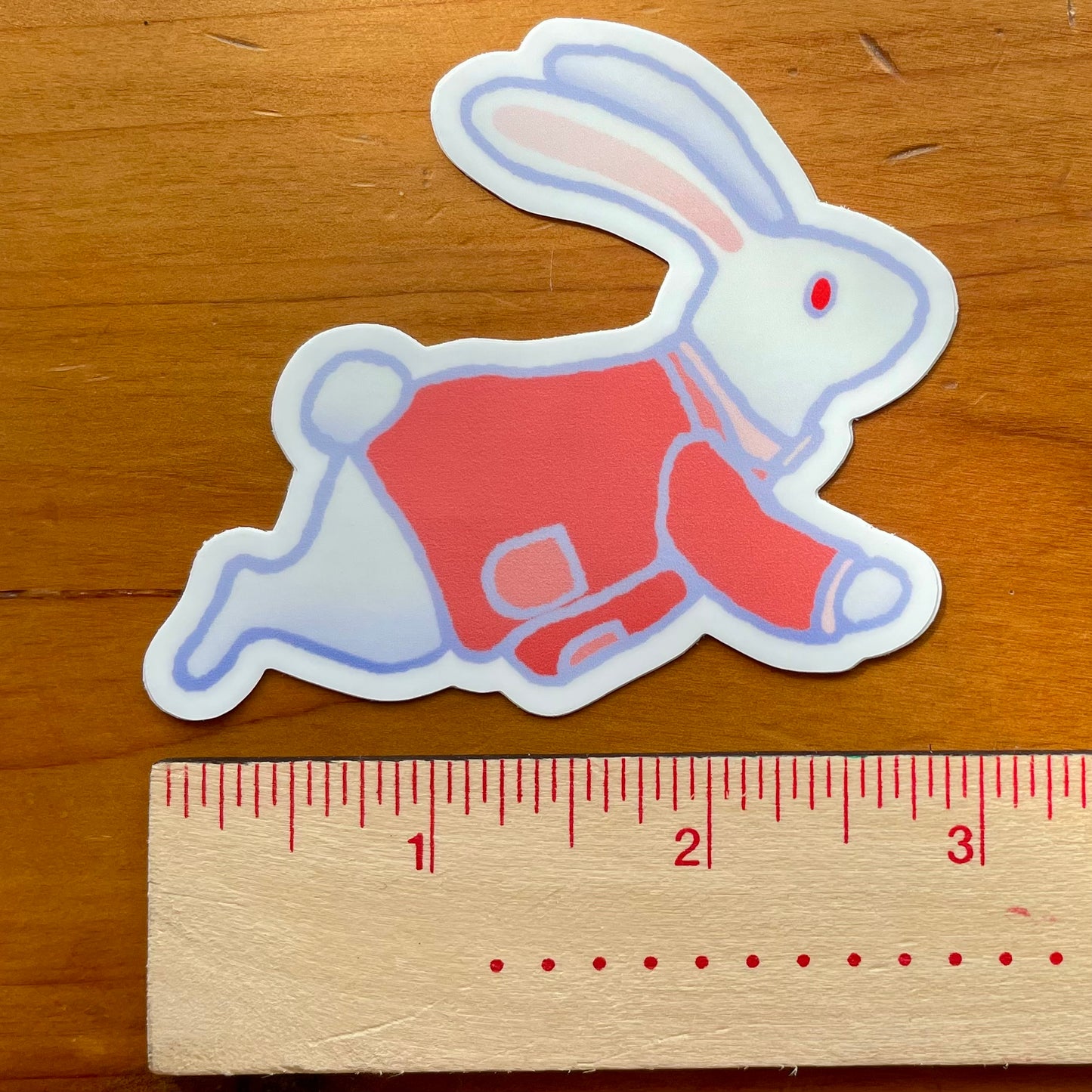 Sticker of The White Rabbit in a red outfit next to a ruler on a wooden surface