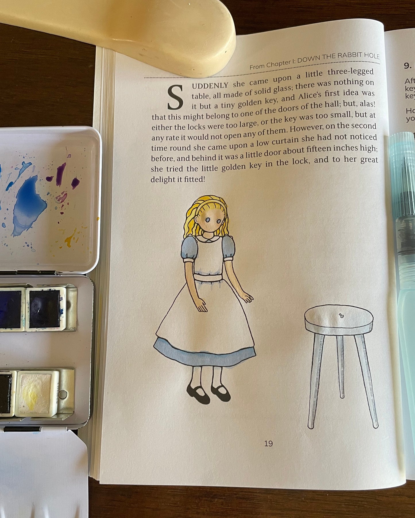Page of The Alice Journal on a wooden table, showing the Alice illustration in watercolor. Surrounding the journal are watercolor palettes and a paintbrush