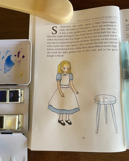 Page of The Alice Journal on a wooden table, showing the Alice illustration in watercolor. Surrounding the journal are watercolor palettes and a paintbrush