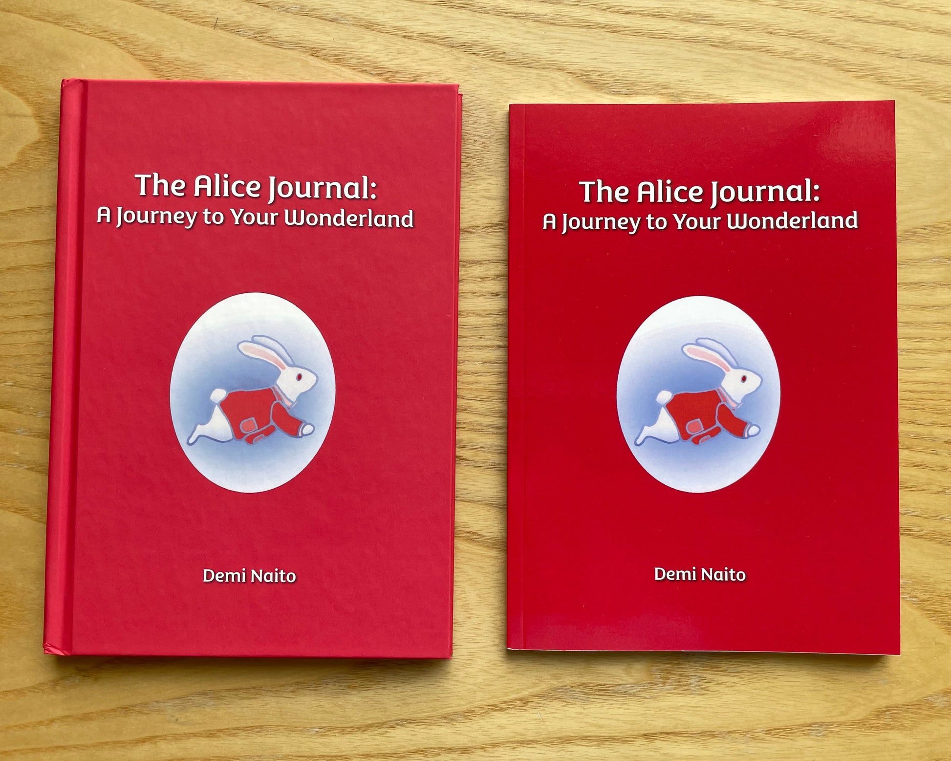 Two copies of the book 'The Alice Journal: A Journey to Your Wonderland' by Demi Naito, featuring red covers with a white rabbit illustration, displayed side-by-side on a light wood background.