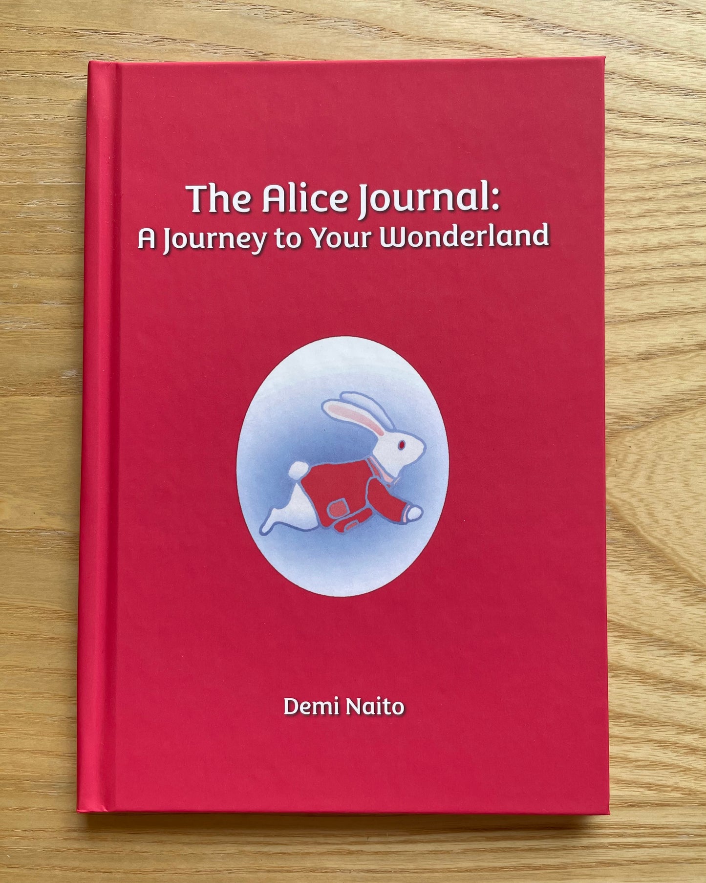 A hardcover journal titled 'The Alice Journal: A Journey to Your Wonderland' by Demi Naito, featuring a bright red cover with a central illustration of a white rabbit in a red outfit inside a light blue oval, placed on a light wood surface.