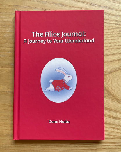 A hardcover journal titled 'The Alice Journal: A Journey to Your Wonderland' by Demi Naito, featuring a bright red cover with a central illustration of a white rabbit in a red outfit inside a light blue oval, placed on a light wood surface.