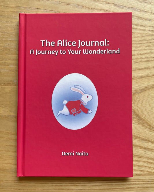 A hardcover journal titled 'The Alice Journal: A Journey to Your Wonderland' by Demi Naito, featuring a bright red cover with a central illustration of a white rabbit in a red outfit inside a light blue oval, placed on a light wood surface.