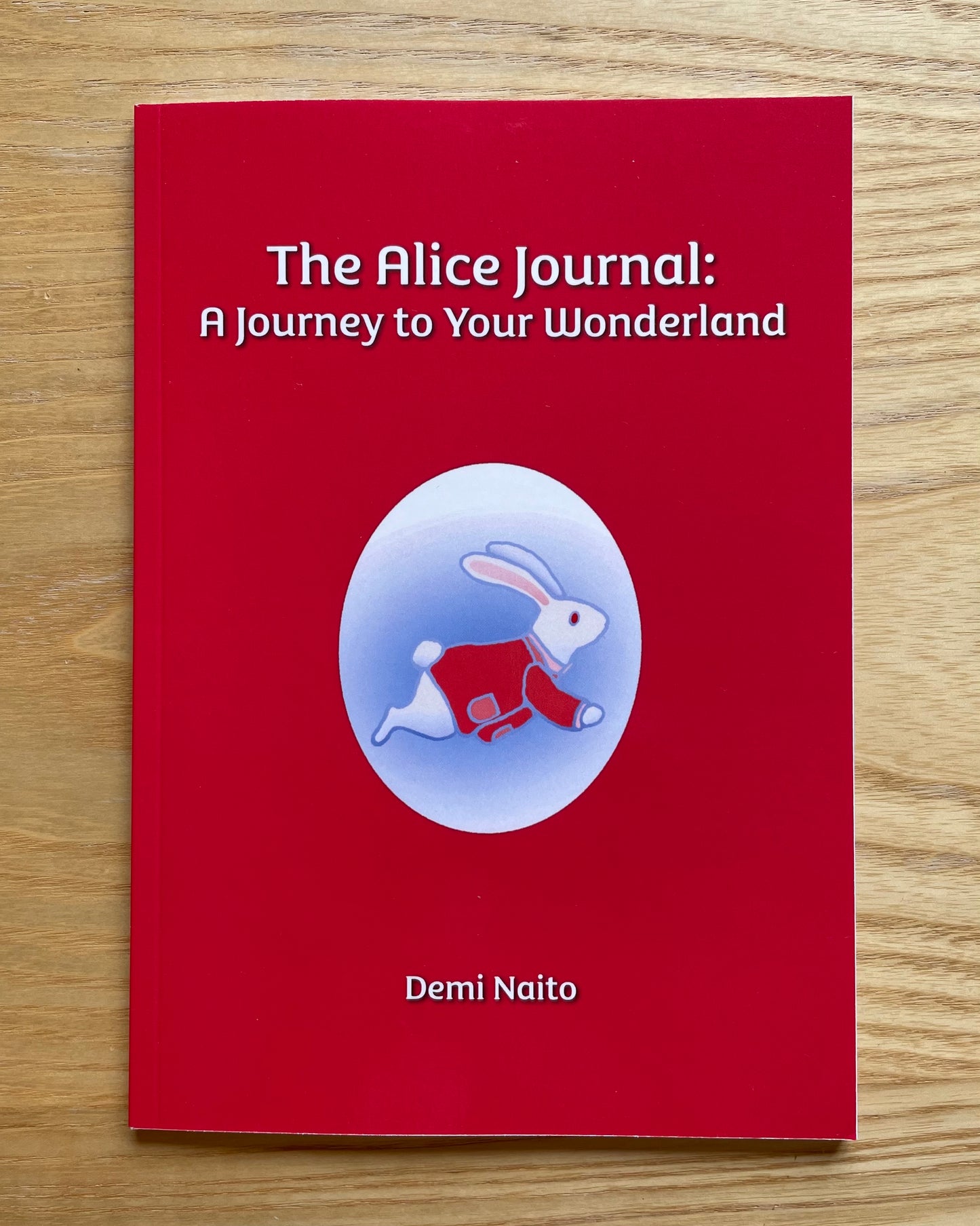 A red hardcover journal titled 'The Alice Journal: A Journey to Your Wonderland' by Demi Naito, featuring a white rabbit illustration in a red outfit inside a light blue oval on the cover, placed on a light wood surface.