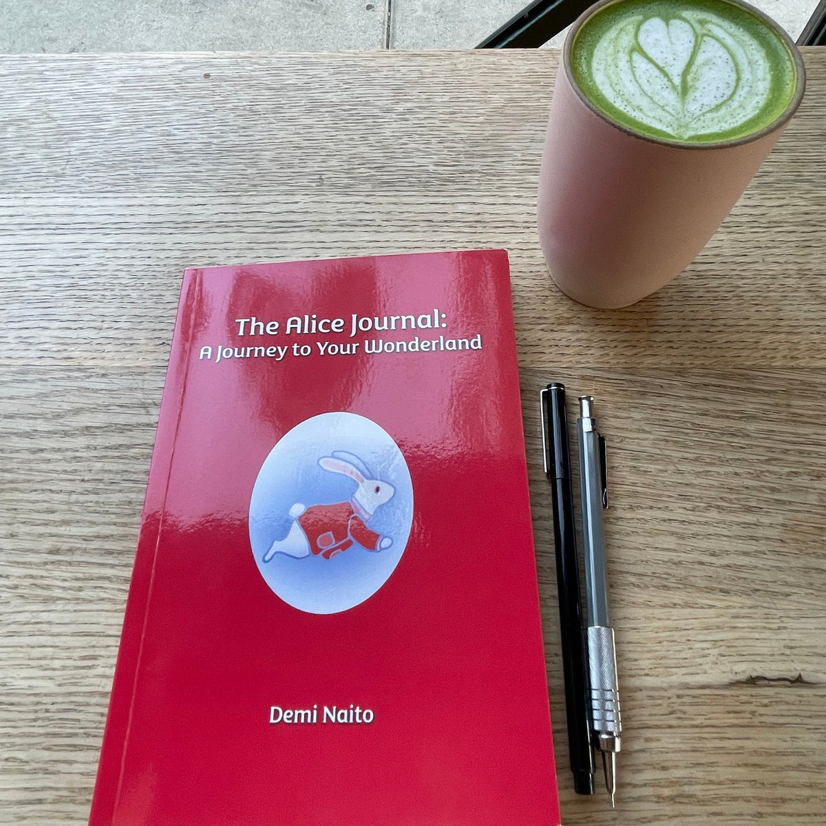 A red softcover book titled 'The Alice Journal: A Journey to Your Wonderland' by Demi Naito lies on a wooden table next to a pink cup of matcha latte with heart-shaped foam art and two black pens.