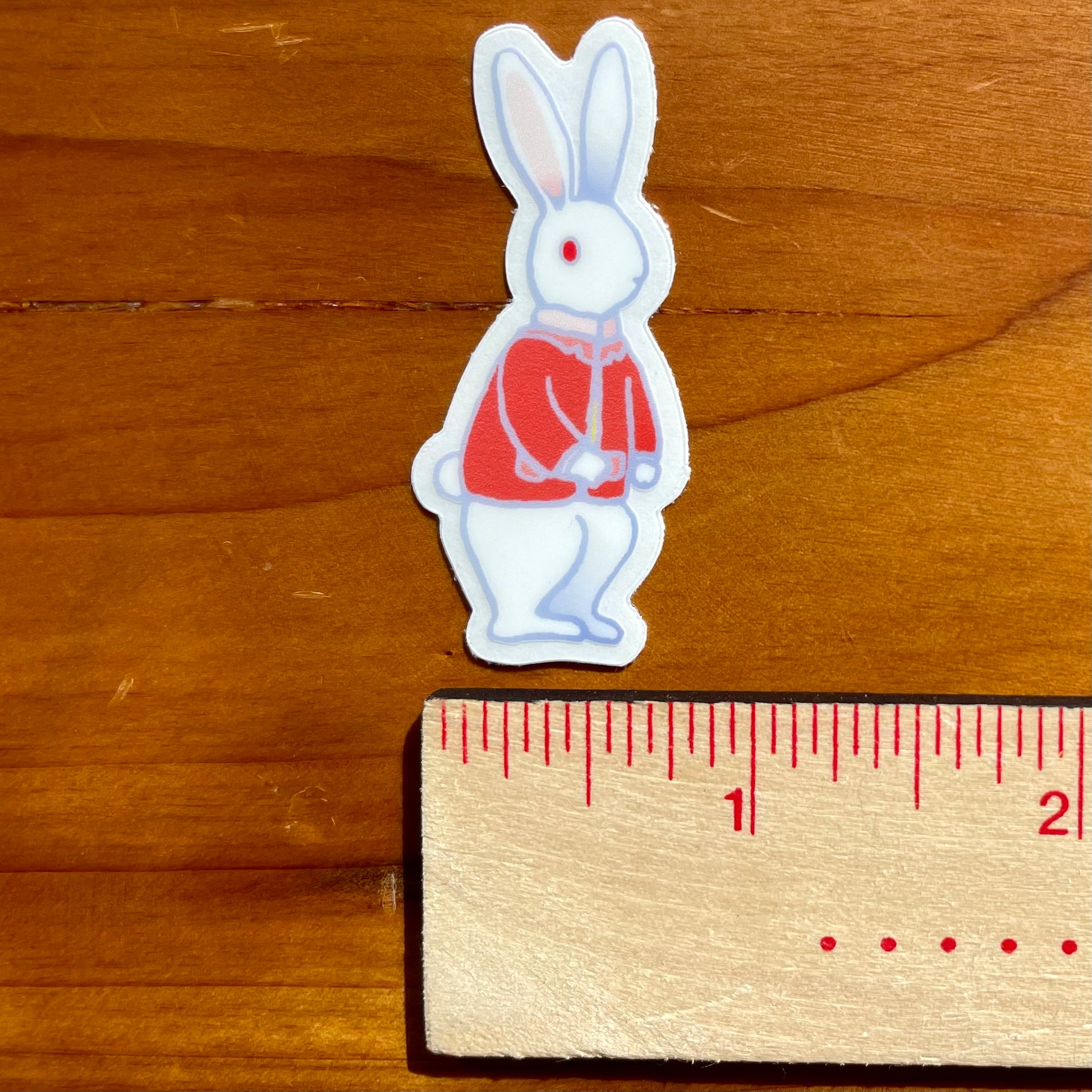 Sticker of a rabbit in a red coat next to a ruler on a wooden surface