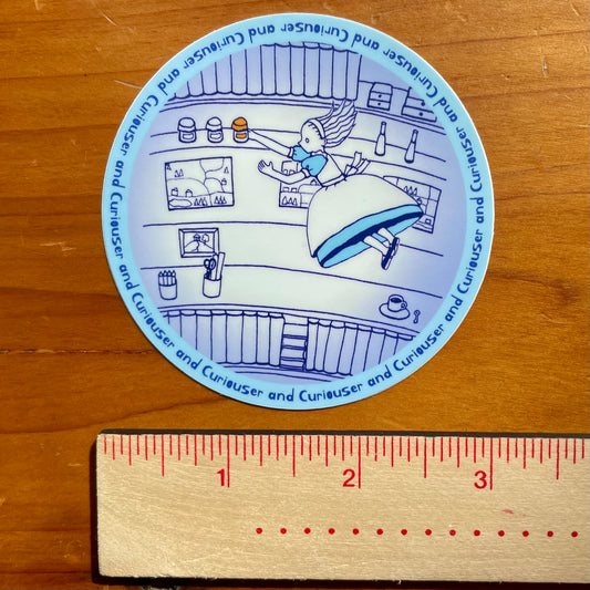 Circular sticker with Alice falling into the well on a wooden surface with a ruler for scale.